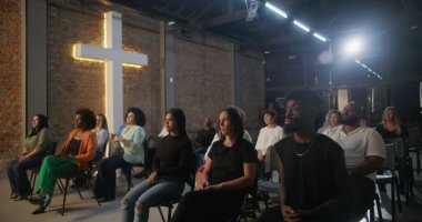 Kilise ayini sırasında oturan cemaat, arka planda aydınlatılmış haçla dikkatle dinliyor, inanç ve ruhsal bağlantı için çeşitli topluluklar bir araya geliyor.