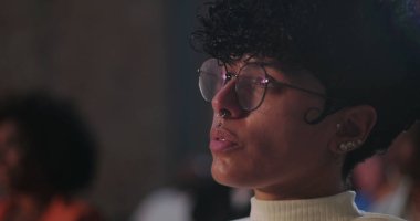 Gözlüklü ve burnu piercingli genç adam dikkatlice uzaklığa bakıyor, yan profil, bir grup ayarında derin odak ve yansıma yakalıyor.