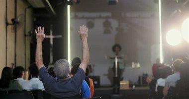 Seyirciler arasında sırtı hoparlöre dönük oturan bir adam, vaaz veren bir kadına dikkat ediyor ya da bir kilise etkinliğinde konuşma yapıyor, arka planda çapraz