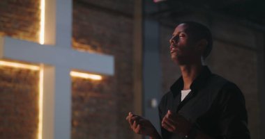 Gözler kapalı dua eden bir adam, dua ederken elleri nazikçe kaldırılmış büyük bir haçın yanında, huzurlu bir yansıma, ruhsal bağlantı ve inanç.