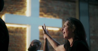 İbadet eden bir kadın, dua ederken gözlerini kapatarak elini kaldırıyor, duygusal bağ kuruyor, Hıristiyan ruhaniliği, tuğla duvarda aydınlatılmış haç, yakınlardaki çeşitli cemaatler.