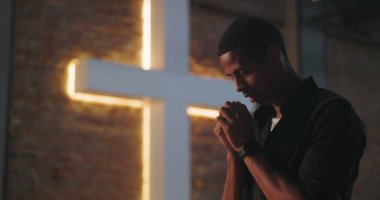 Aydınlatılmış haçın yanında ellerini sımsıkı bağlayıp dua eden adam, aşağıya bakıyor, huzur dolu ruhsal yansıma, Hristiyan inancı, ibadet ve duaya olan güven.