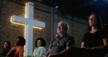 Arka planda aydınlanmış haçla kilisede oturan çeşitli insan grupları, ibadet sırasında maneviyat ve sadakat, birlik ve dini cemaatle bağlantıyı ifade ediyorlar.