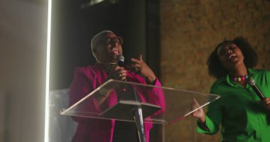 Afrika kökenli kadınlar tutkulu bir şekilde kilise ibadeti sırasında şarkı söylüyorlar, mikrofon tutuyorlar, anlamlı jestler yapıyorlar, cemaate içten dini bir performans sergiliyorlar.