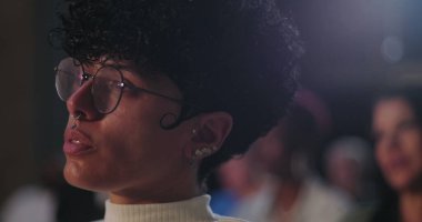 Gözlüklü ve burnu piercingli genç adam dikkatlice uzaklığa bakıyor, yan profil, bir grup ayarında derin odak ve yansıma yakalıyor.