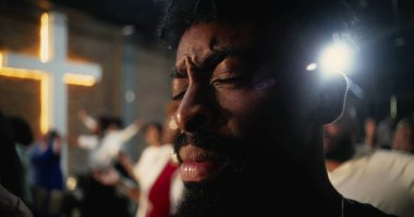 İbadet sırasında dua eden Afrikalı bir Amerikalının yakın çekimi, gözleri derin inanç ve adanmışlıkla kapalı, arka planda aydınlanan haç ruhaniliği ve umudu simgeliyor.