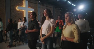 Arka planda parlayan bir haçla Hıristiyan kilisesi etkinliğine katılan çeşitli insanlar ellerini kaldırarak ibadet ediyorlar.