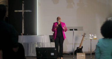 Vaaz veren, mikrofon tutan, arka planda haç olan kilisedeki cemaate hitap eden, dinleyicilerin el çırpması, Hıristiyan ruhani buluşması.