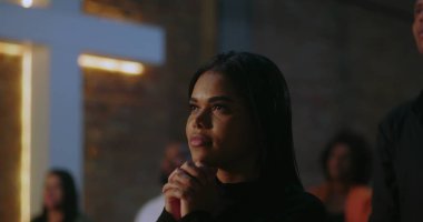 Elleri sımsıkı bağlı dua eden genç bir kadın, inanç ve umutla gözlerini kaldırmış, kilisede aydınlatılmış haçın önünde dikilmiş, arka planda birbirine tapan bir cemaat.