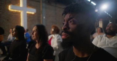 Afro-Amerikalı bir adamın kilise ayini sırasında gülümseyen, neşe ve bağlantıyı ifade eden arka planda görünen tuğla duvarda yanan bir haç profili.