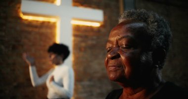 Kilise ayini sırasında namaz kılan yaşlı bir kadın, elleri göğsünde, gözleri bağlılık içinde kapalı, arka planda aydınlanmış haç ve tuğla duvar