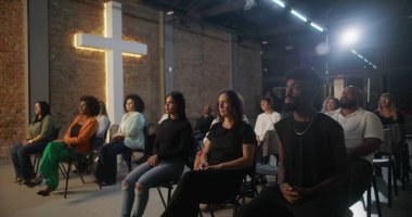 Kilise ayini sırasında oturan cemaat, arka planda aydınlatılmış haçla dikkatle dinliyor, inanç ve ruhsal bağlantı için çeşitli topluluklar bir araya geliyor.