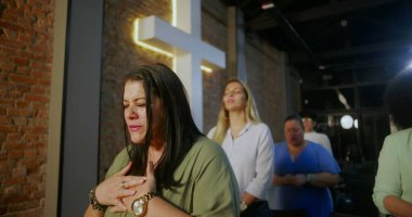 Kilisenin içinde elleri üzerinde dua eden bir kadın, derin bir bağlılıkla gözlerini kapatmış, diğer ibadet edenler tarafından çevrelenmiş, ve tuğla bir duvarda parlayan bir haç