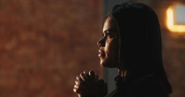 Elleri sımsıkı bağlı dua eden genç bir kadının yan profili, inancın huzurlu ifadesi, yumuşak bir ışık, arka planda dokulu tuğla bir duvar, ruhani bir bağlılık.