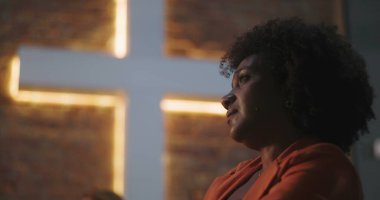 Kıvırcık saçlı bir kadın, kilisede aydınlanmış bir haçın önünde derin bir ibadete dalmış, ruhani bağlantı, inanç, tefekkür, dua anı.
