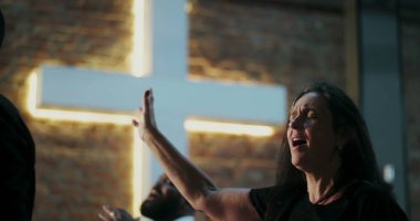 İbadet eden bir kadın, dua ederken gözlerini kapatarak elini kaldırıyor, duygusal bağ kuruyor, Hıristiyan ruhaniliği, tuğla duvarda aydınlatılmış haç, yakınlardaki çeşitli cemaatler.