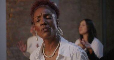 Huzurlu bir ifadeyle şarkı söyleyen bir kadın, bir kilise ayini sırasında ruhaniliğe ve inanca dalmış, arka planda aydınlatılmış bir haç