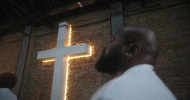 Kilise ayinine katılan kişi, modern kilise ortamında aydınlanmış haçın yanında ruhani bir yansımayla yukarı bakıyor, çeşitli cemaatlerde ibadet ediyor.