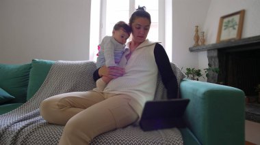 Anne kanepede bebeği tutuyor, ikisi de tablete bakıyor, birlikte ekran başında vakit geçiriyorlar, sıcak bir oturma odasında rahat bir aile zamanı geçiriyorlar..