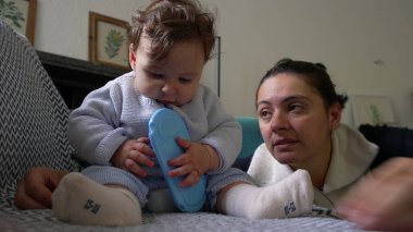 Bebek koltukta oturup mavi nesneyi çiğniyor, anne sevgiyle izliyor, kapalı alanda oyun oynuyor, bebek keşif yapıyor, aile bağı kuruyor, rahatlamış ev ortamları, çocuk gelişimi..