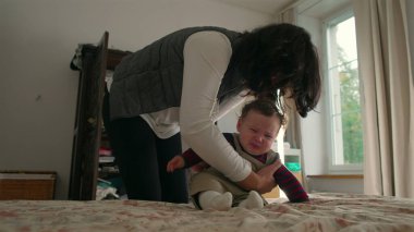 Anne bebek elbiselerini ayarlıyor ve onu yataktan kaldırıyor, bebek sırtlarını ve kollarını havada destekliyor, sakin bir evde sabah rutinini besliyor..