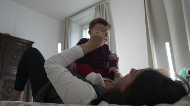 Anne bebekle yatakta uzanıyor, nazikçe oynuyor ve çocukla bağ kuruyor, erken duygusal gelişimi ve sıcak bir ev ortamında bağlantıyı besliyor..