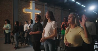 Arka planda parlayan bir haçla Hıristiyan kilisesi etkinliğine katılan çeşitli insanlar ellerini kaldırarak ibadet ediyorlar.