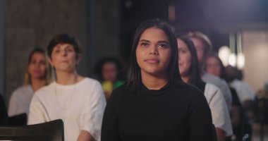 Bir grup insan, derin düşüncelere dalmış, ışıkla aydınlatılmış, diğerleri ise arka planda bulanık bir kadına odaklanmış bir etkinliğe katılıyorlar.