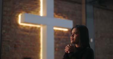İspanyol bir kadının elleri sımsıkı bağlı, yumuşak bir ifadeyle dua edişi, evanjelist ibadet sırasında arka planda ışıklandırılmış haç, umudu, maneviyatı ve inancı sembolize ediyor.