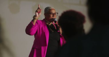 Sözcü tutkulu bir şekilde dinleyicilere hitap ediyor, mikrofonu tutuyor, vurgu için elini işaret ediyor, güçlü vaaz dağıtımı, çeşitli cemaatler dikkatlice dinliyor, ruhani olay