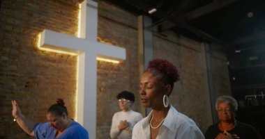 Kilisenin içinde dua eden bir kadın, minnettarlığını ve inancını ifade eden, arka planda diğer ibadet edenlerle birlikte ışıldayan bir haçın önünde duran.