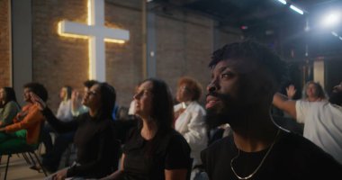 Kilise ayini sırasında şarkı söyleyen ve ibadet eden çeşitli cemaatler, arka planda aydınlatılmış haç ve tuğla duvar, maneviyat ve inancı ifade ediyor.