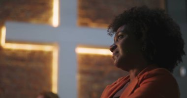 Kıvırcık saçlı bir kadın, kilisede aydınlanmış bir haçın önünde derin bir ibadete dalmış, ruhani bağlantı, inanç, tefekkür, dua anı.