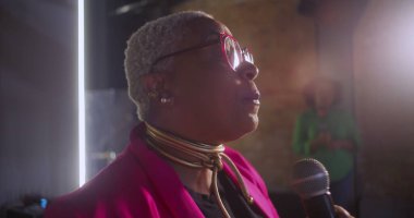 Bir kadın konuşmacının tutkuyla vaaz verirken, ifade ve bağlantıya odaklı bir mikrofonu tutarken, arka planda bulanık duran tuğla duvar ve katılımcının yakın plan görüntüleri.