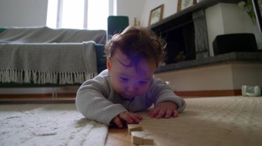 Bebek yerde yatıyor, ahşap bir bloğu yakından inceliyor, merakla eğiliyor, etkin bir şekilde çevreyi keşfediyor, erken çocukluk öğrenimi, rahat bir evde dokunsal bir keşif..