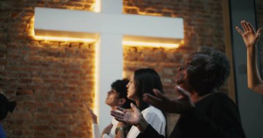 Kilisede el kaldırarak ibadet eden çeşitli gruplar, birliğe olan inanç ve bağlılıklarını ifade eden, arka planda aydınlanan haç ruhaniliği simgeliyor.