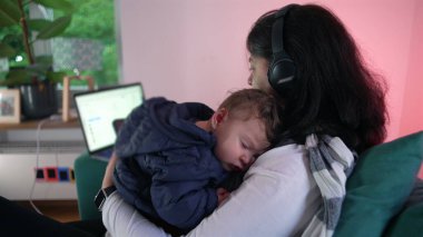 Anne kucağında bir dizüstü bilgisayar, rahat bir ev atmosferinde iş ve anneliği birleştirerek kariyer ve bakım sorumluluklarını dengeliyor..