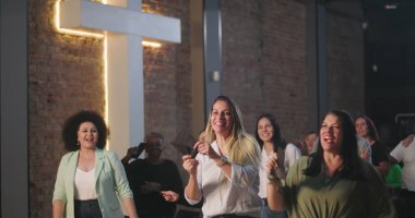 Kiliseye ibadet sırasında bağış kutusuna zarf koyan iki kadın, neşeli ve saygılı ifadeler, ruhani sunuma katılan çeşitli arka planda yer alan farklı topluluk