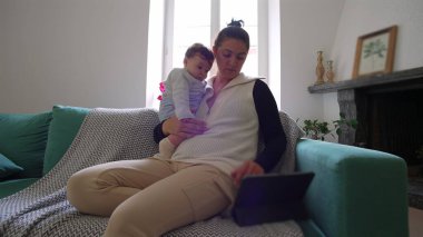 Anne kanepede bebeği tutuyor, ikisi de tablete bakıyor, birlikte ekran başında vakit geçiriyorlar, sıcak bir oturma odasında rahat bir aile zamanı geçiriyorlar..