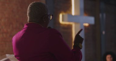 Afrikalı Amerikalı vaiz, arka planda aydınlanmış haçla mesajını iletiyor. Kutsal kilise ortamında güçlü bir liderlik ve inanç anı. Ruhani bağlantı vurgulanıyor.