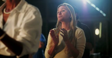 Manevi bir buluşmada el çırpan kadın, düşünceli ifade, ibadet ortamında çeşitli insanlarla çevrili, sadakat, inanç ve topluluk bağlantısına odaklanmış.