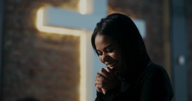 Evanjelist ibadet sırasında gülümseyen İspanyol kadının yakın çekimi, el ele tutuşmuş bağlılık, derin inanç ve ruhaniliği simgeleyen arka planda aydınlatılmış haç.