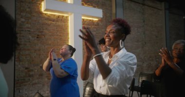 Cemaat kilise ayini sırasında el çırpıyor, arka planda haç ve tuğla duvarlar aydınlatılıyor, çeşitli topluluklar neşe ve birlik içinde.