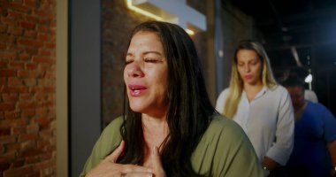 Kilisenin içinde elleri üzerinde dua eden bir kadın, derin bir bağlılıkla gözlerini kapatmış, diğer ibadet edenler tarafından çevrelenmiş, ve tuğla bir duvarda parlayan bir haç