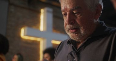 Kilisede namaz kılarken yansıyan yaşlı adamın yakın çekimi, sadakat içinde sıkıca sarılmış eller, arka planda aydınlatılmış haç, derin inanç ve ruhaniliğin ifadesi.