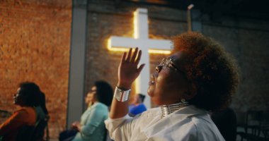 İbadet sırasında dua eden Afrikalı Amerikalı bir kadın. Gözler derin ruhsal bağlantıda kapalı. Arka plandaki haç ve cemaat aydınlanmış.