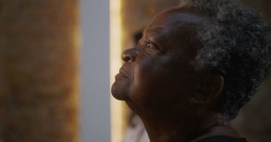 Kilisede ibadet ederken sakin bir ifadeyle profil görünümlü yaşlı bir kadın, arka planda aydınlanmış haç, maneviyat, inanç ve odak noktasında toplum