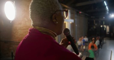 Konuşmacı mikrofonu tutarken vaaz veriyor, tutkulu bir şekilde işaret ediyor, arka plandaki tuğla duvarda yanan haç, dinleyicileri inanç ve ruhsal bağlantıyla meşgul ediyor.