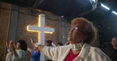 İbadet sırasında el ele tutuşan, birlik ve inanç içinde duran, arka planda toplumu ve ruhsal bağlantıyı simgeleyen haçı aydınlatan cemaat.