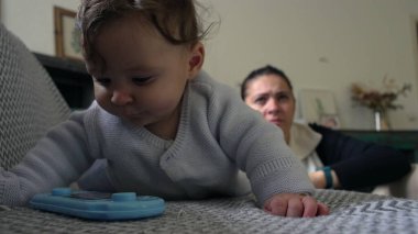 Bebek koltuğa yaslanırken mavi nesneyle oynuyor, anne arkadan izliyor, bebek keşfe çıkıyor, eğlenceli bir öğrenme anı, evde anne ile vakit geçiriyor..